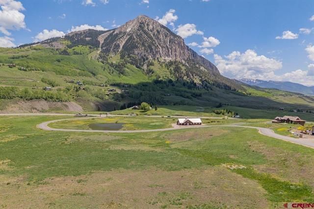 Mountain landscape with green fields, a winding dirt road, and a small white farmhouse under a blue sky.