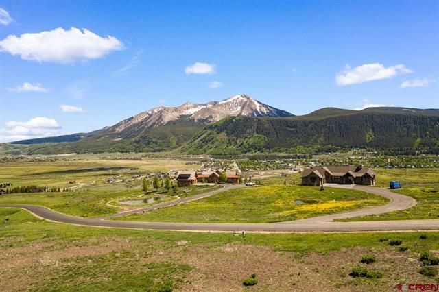Rural mountain landscape with a winding road, green fields, and small cabins under a blue sky