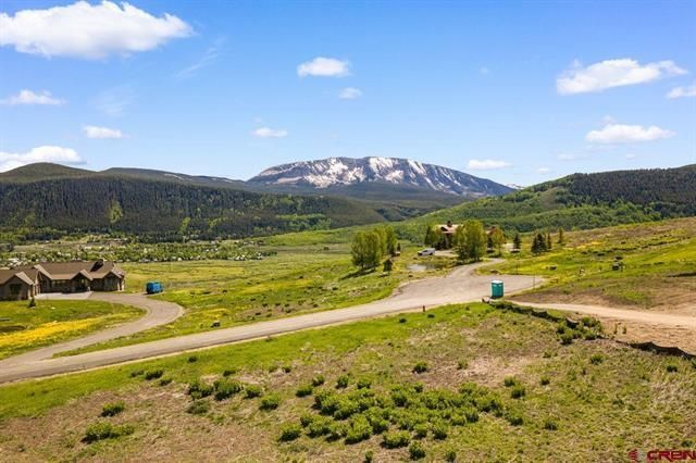 Mountain landscape with green meadow, winding dirt road, and snow-capped peak under a blue sky