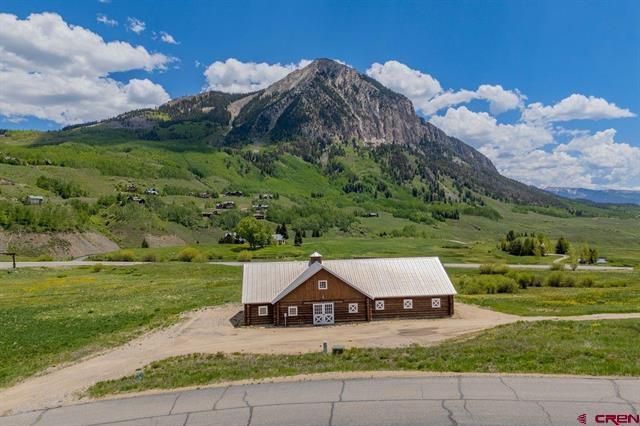 Small red cabin in green valley below a rocky mountain under a blue sky with clouds