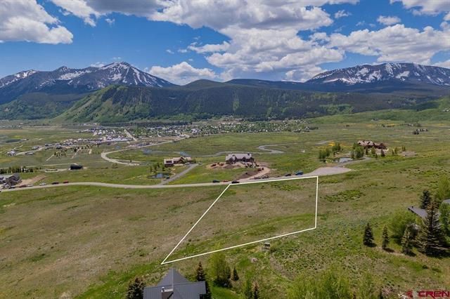 Mountain valley town with open fields, roads, and snow-capped peaks under a blue sky