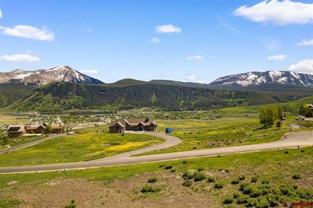 Mountain valley with winding road, green fields, scattered houses, and snow-capped peaks under a blue sky