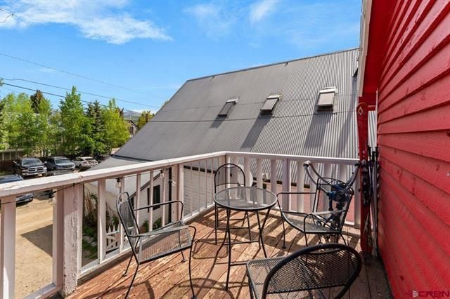 Balcony with metal chairs and table overlooking a red-sided building and gray-roofed structure under blue sky