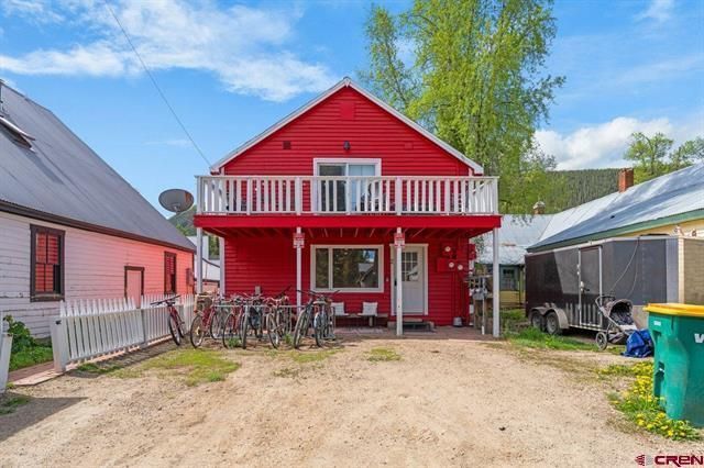 Bright red two-story house with a balcony, bicycles in front, and a dirt driveway on a sunny day