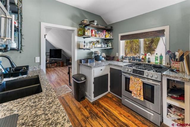 Small kitchen with stainless stove, granite counters, shelves, and hardwood floors leading to a living room.