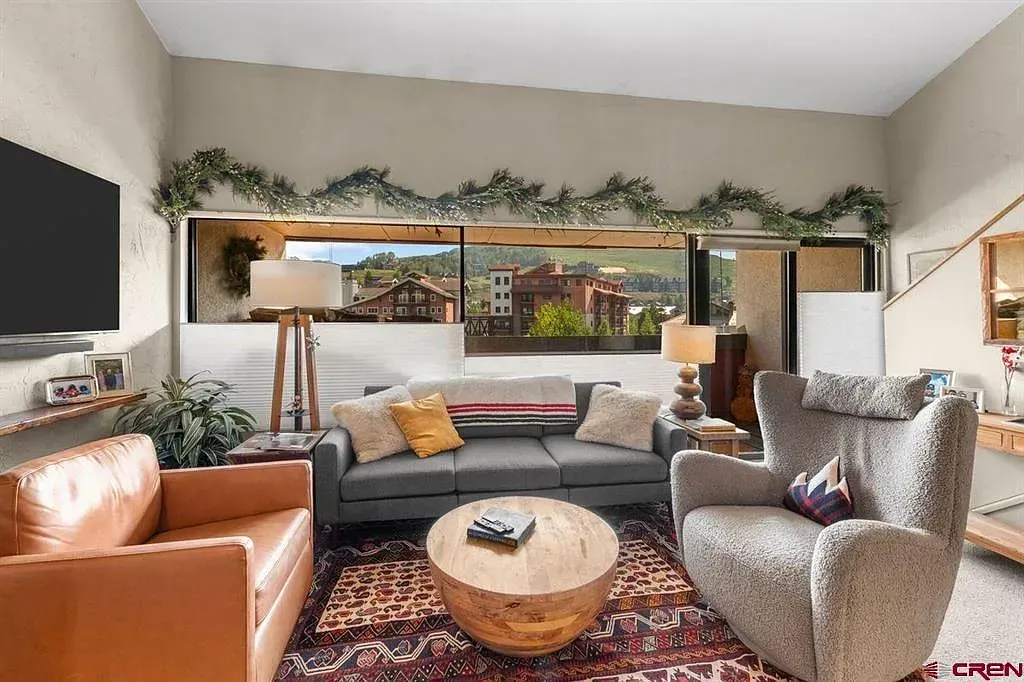 Cozy living room with gray sectional, tan leather chair, round wood coffee table, and large windows.