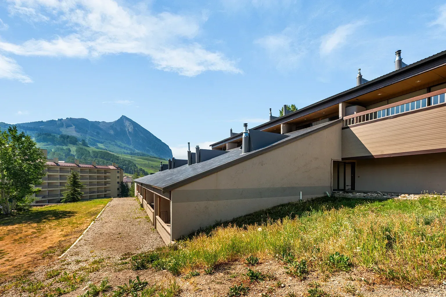 Sunny mountain-side building with beige walls, grassy slope, and distant peaks under a blue sky