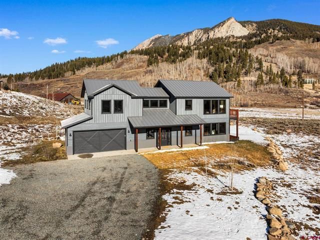 Gray two-story house with mountain backdrop and patchy snow in a rural setting