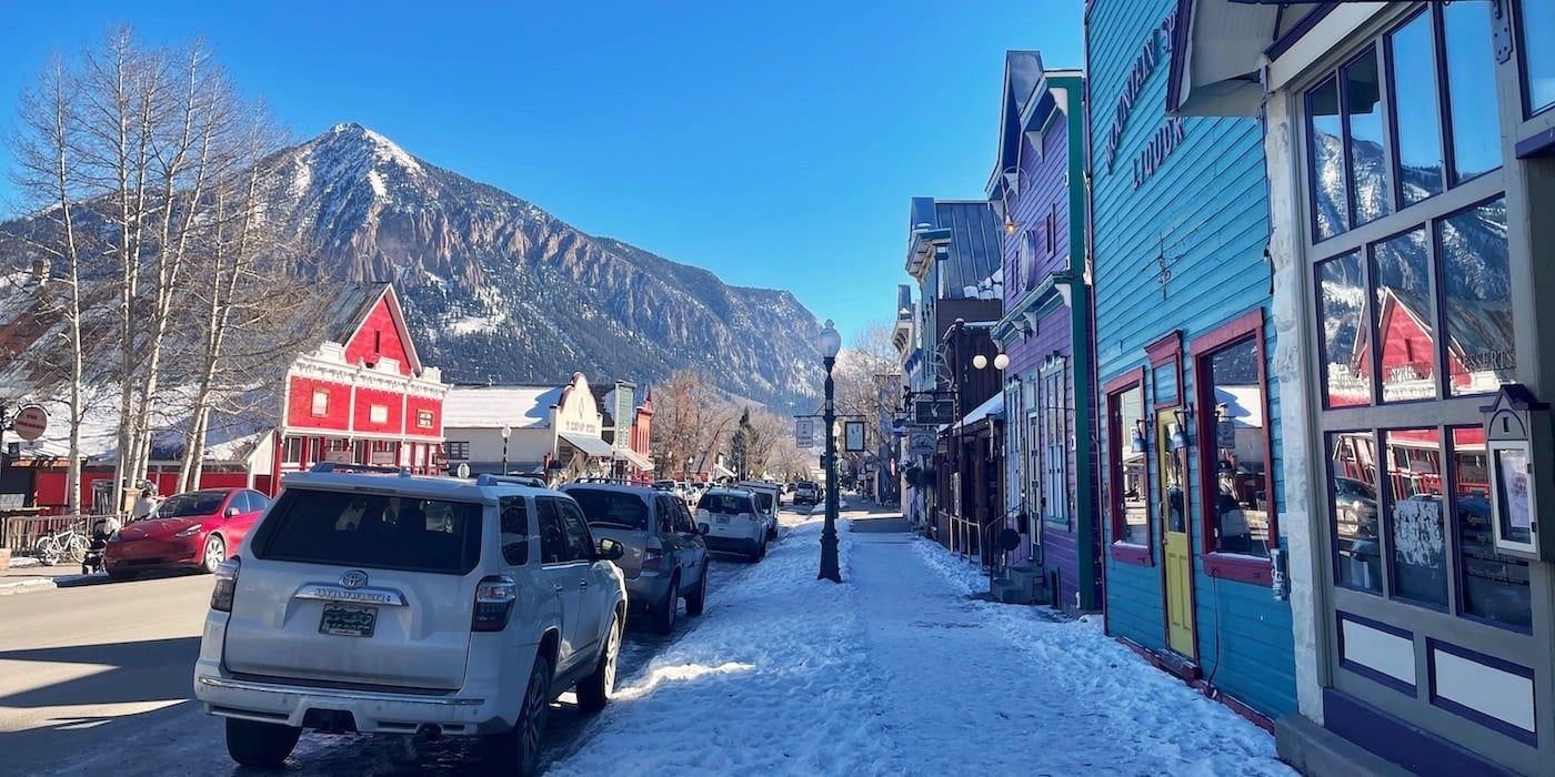 Snowy mountain town street with parked cars, colorful storefronts, and a clear blue sky