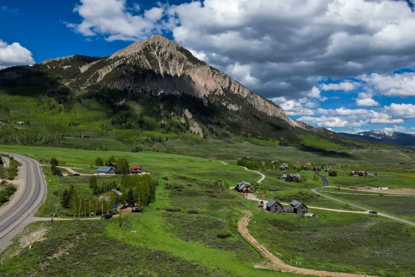 Green valley with a winding road, scattered houses, and a rocky mountain under dramatic clouds