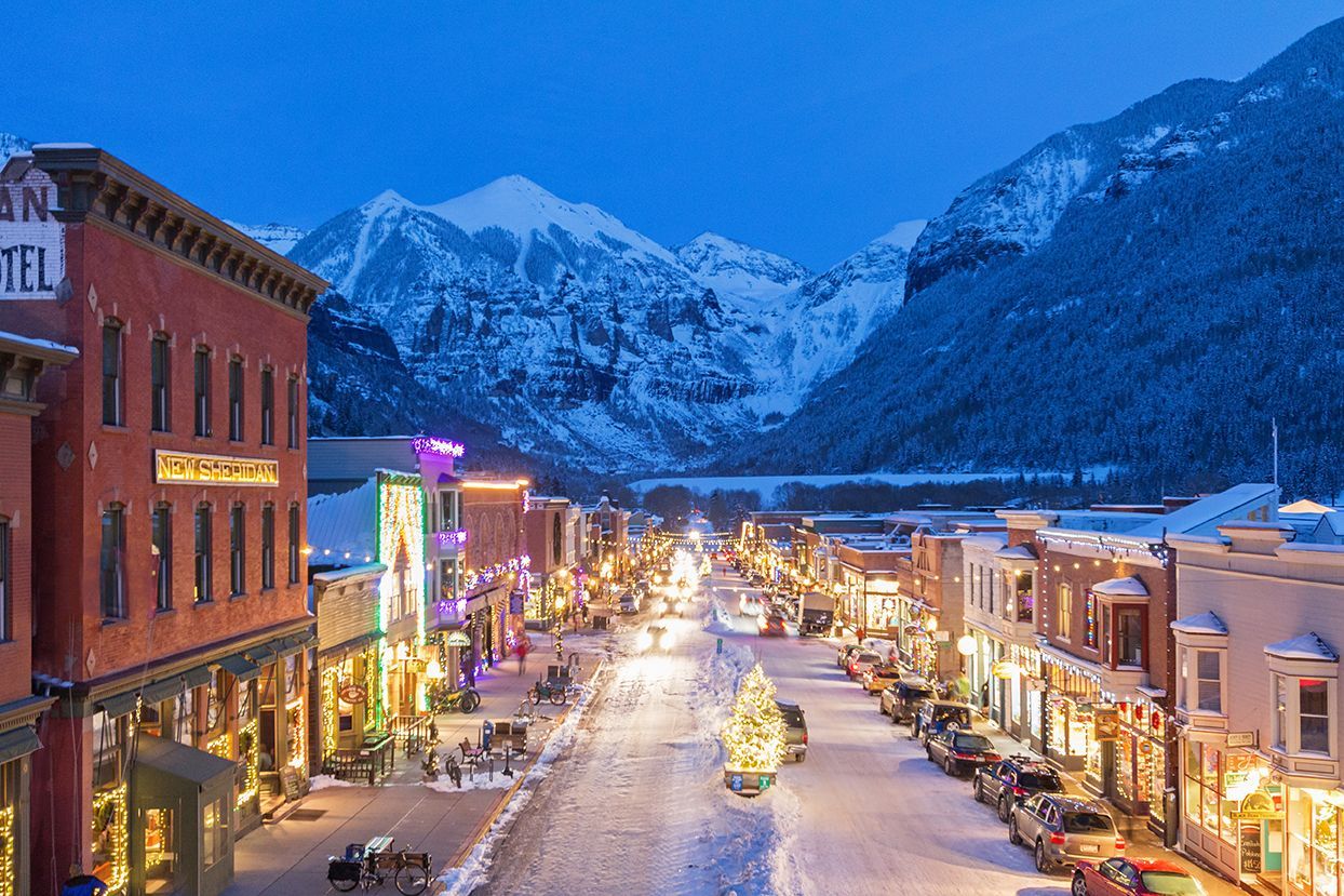 Snowy mountain town street at dusk, lit with festive holiday lights and storefronts under blue twilight.