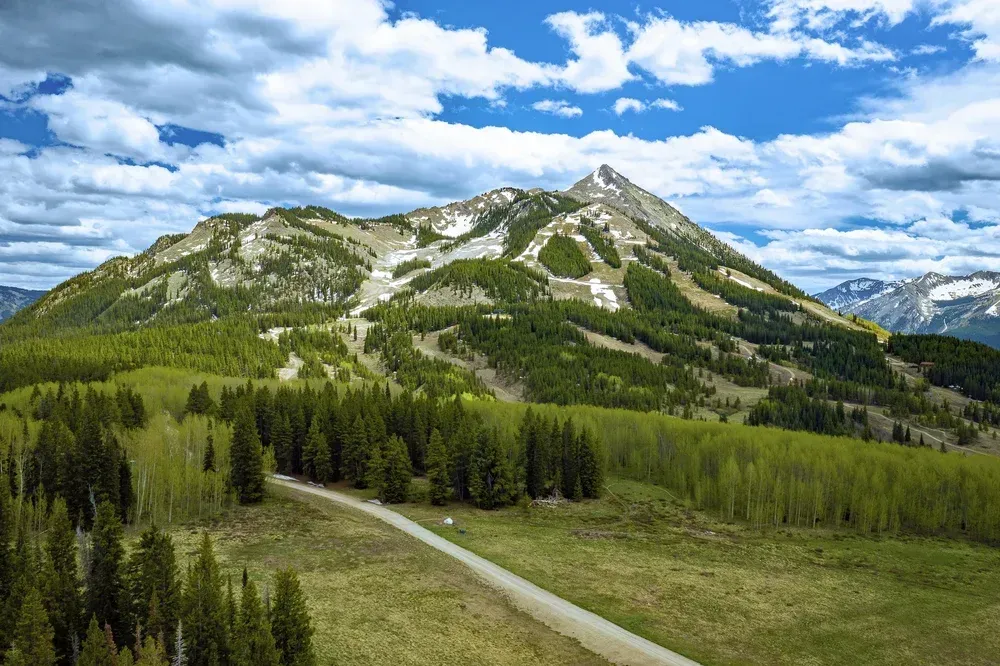 Mountain landscape with green forest, winding road, and rocky peak under a blue sky with clouds