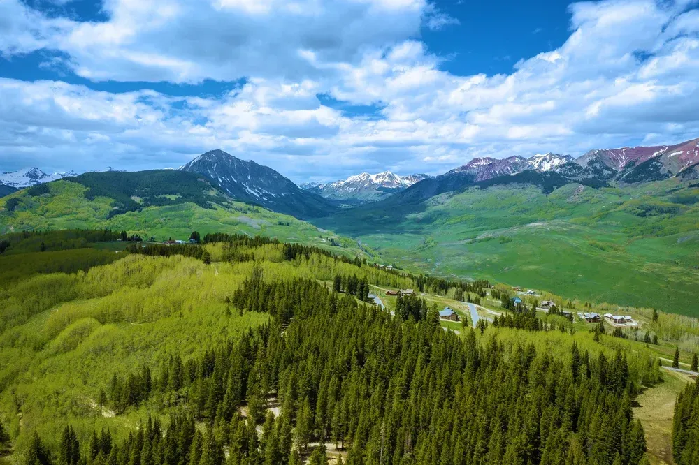Green mountain valley with dense forest, winding road, and snow-capped peaks under a cloudy blue sky