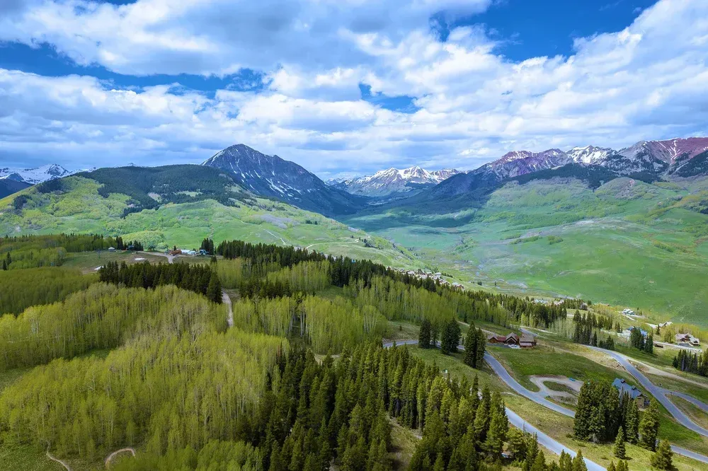 Green alpine valley with winding road, forested hills, and snow-capped mountains under a cloudy blue sky
