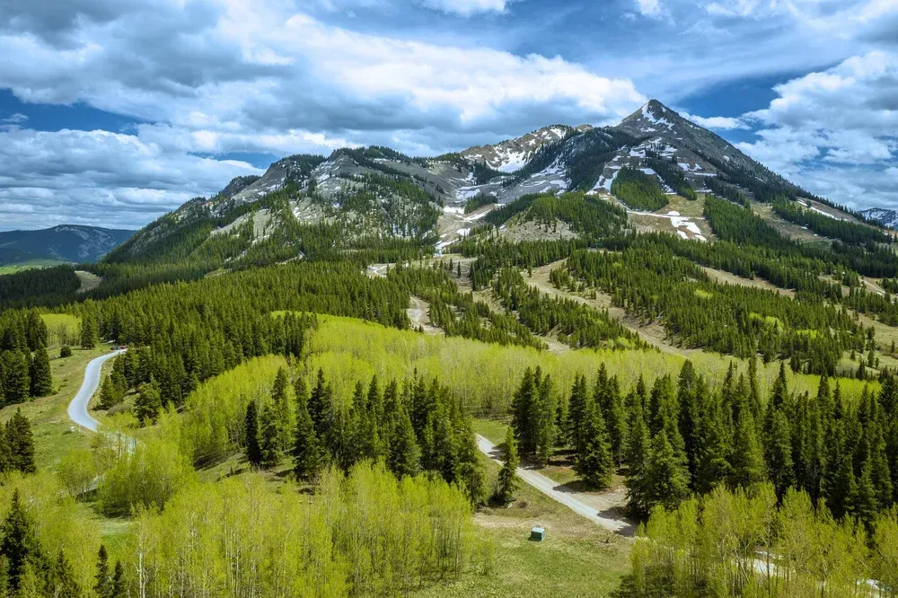 Mountain landscape with winding road, green forest, and rocky peaks under a cloudy sky