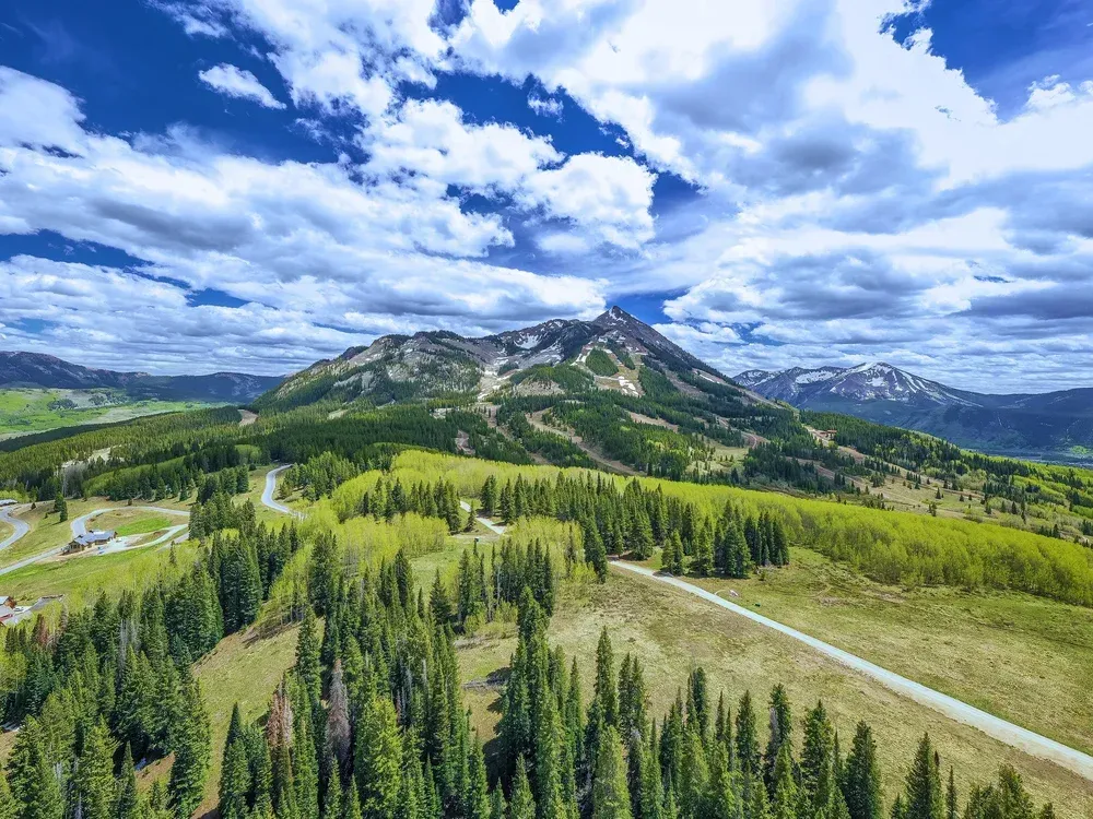 Mountain landscape with green forests, winding road, and dramatic clouds over snowy peaks