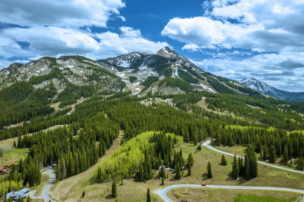 Mountain landscape with evergreen forests, rocky peaks, and a winding road under a bright blue sky