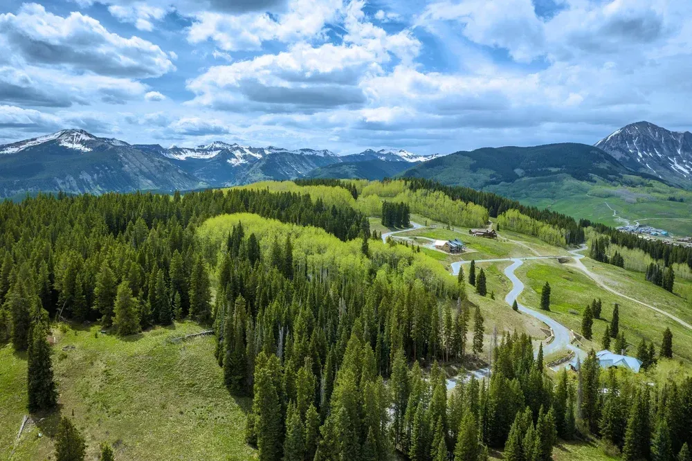 Mountain landscape with green forested hills, a winding road, and snow-capped peaks under cloudy sky