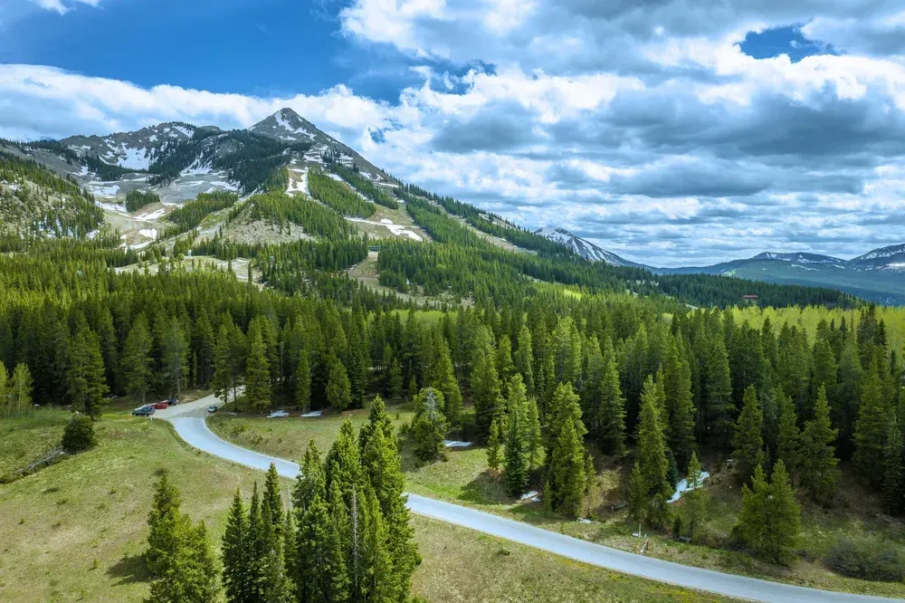 Mountain landscape with winding road, evergreen forest, and snow-dusted peaks under cloudy blue sky