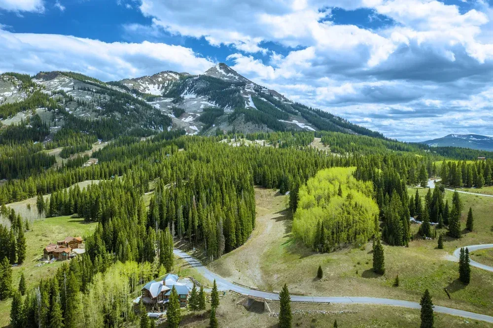 Mountain valley with dense evergreen forest, winding creek, and snow-dusted peaks under a cloudy blue sky