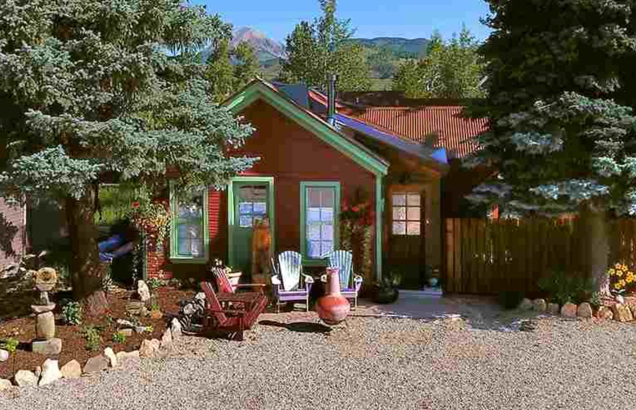 Rustic cabin with red roof, gravel yard, chairs, and potted plants amid trees and mountains