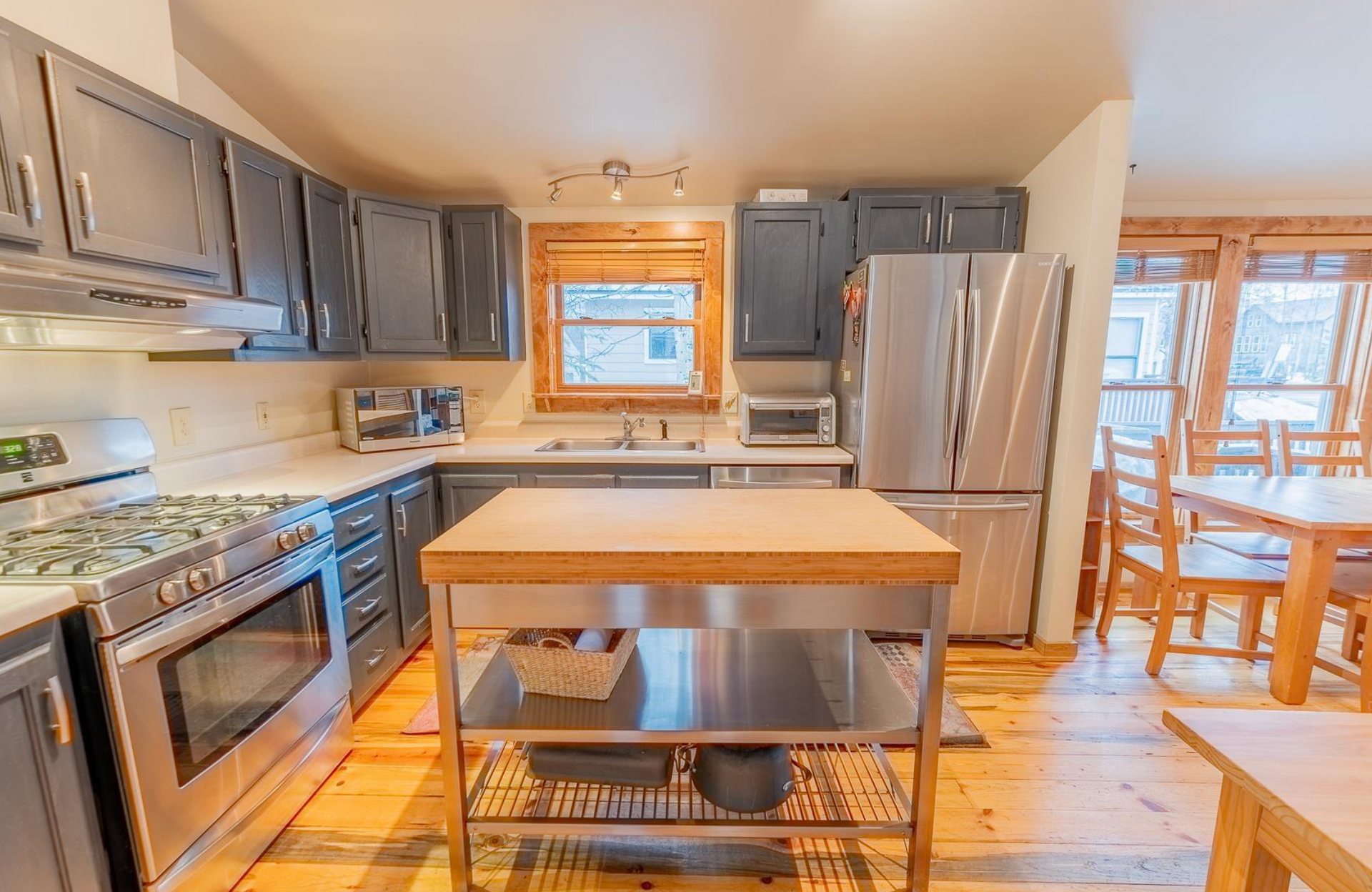 Bright kitchen with gray cabinets, stainless appliances, wood island, and dining area by a window.