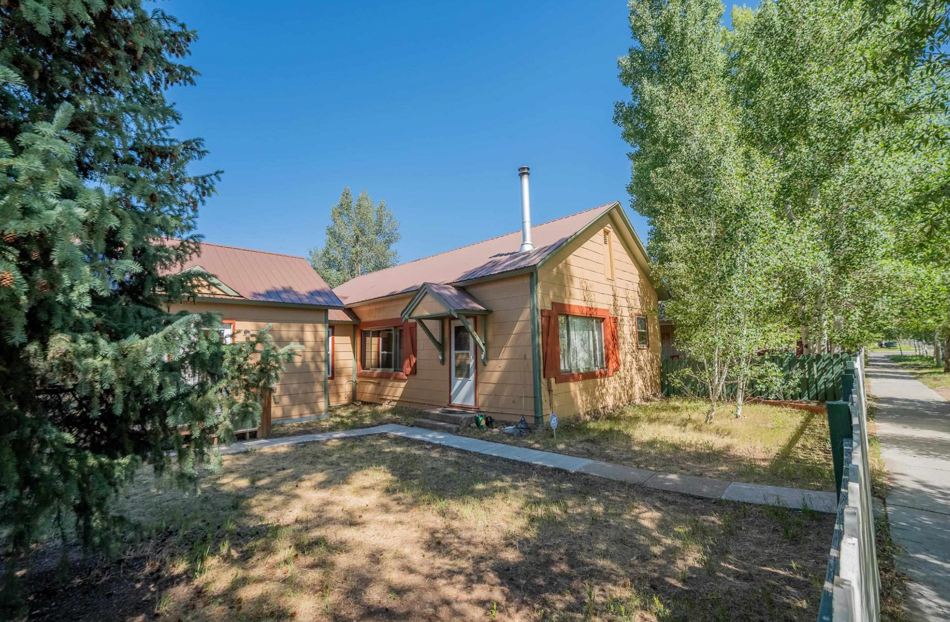 Small tan house with red shutters and a fenced yard under a clear blue sky.