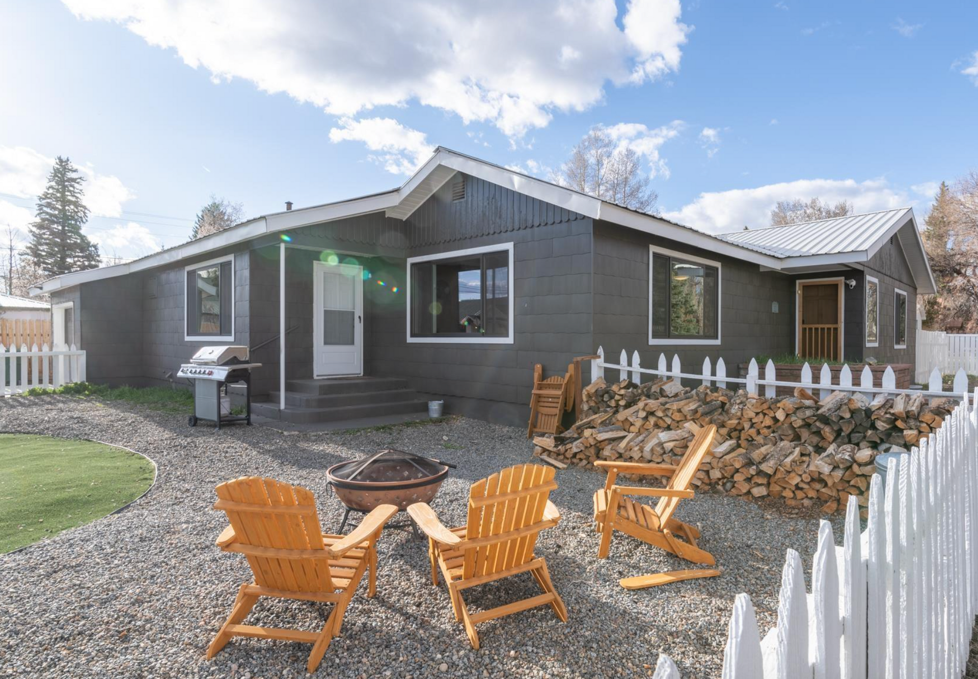 Modern gray house with a gravel patio, orange chairs, and white picket fence under a partly cloudy sky
