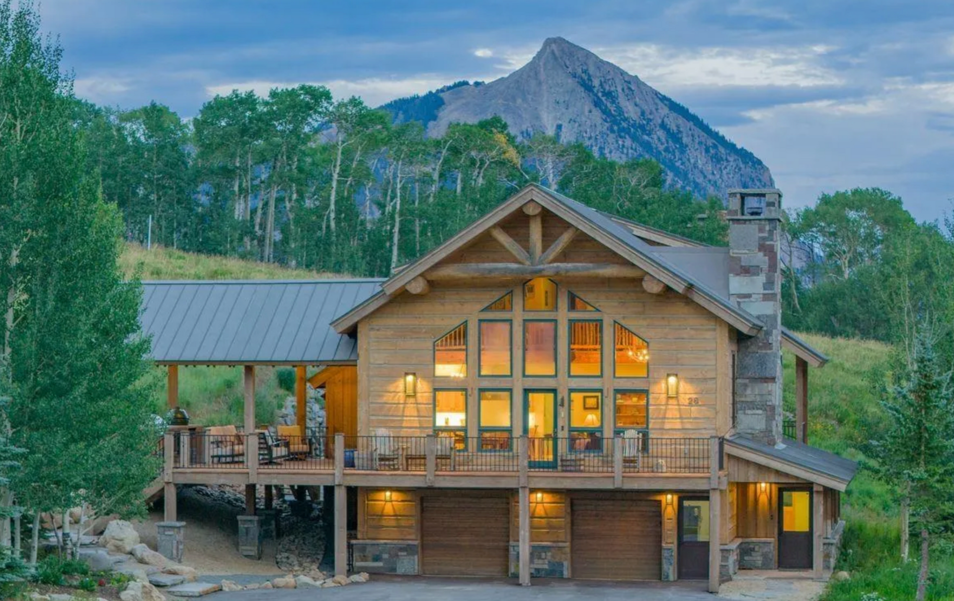 Rustic mountain lodge with warm lights and a stone chimney, set among trees beneath a rocky peak