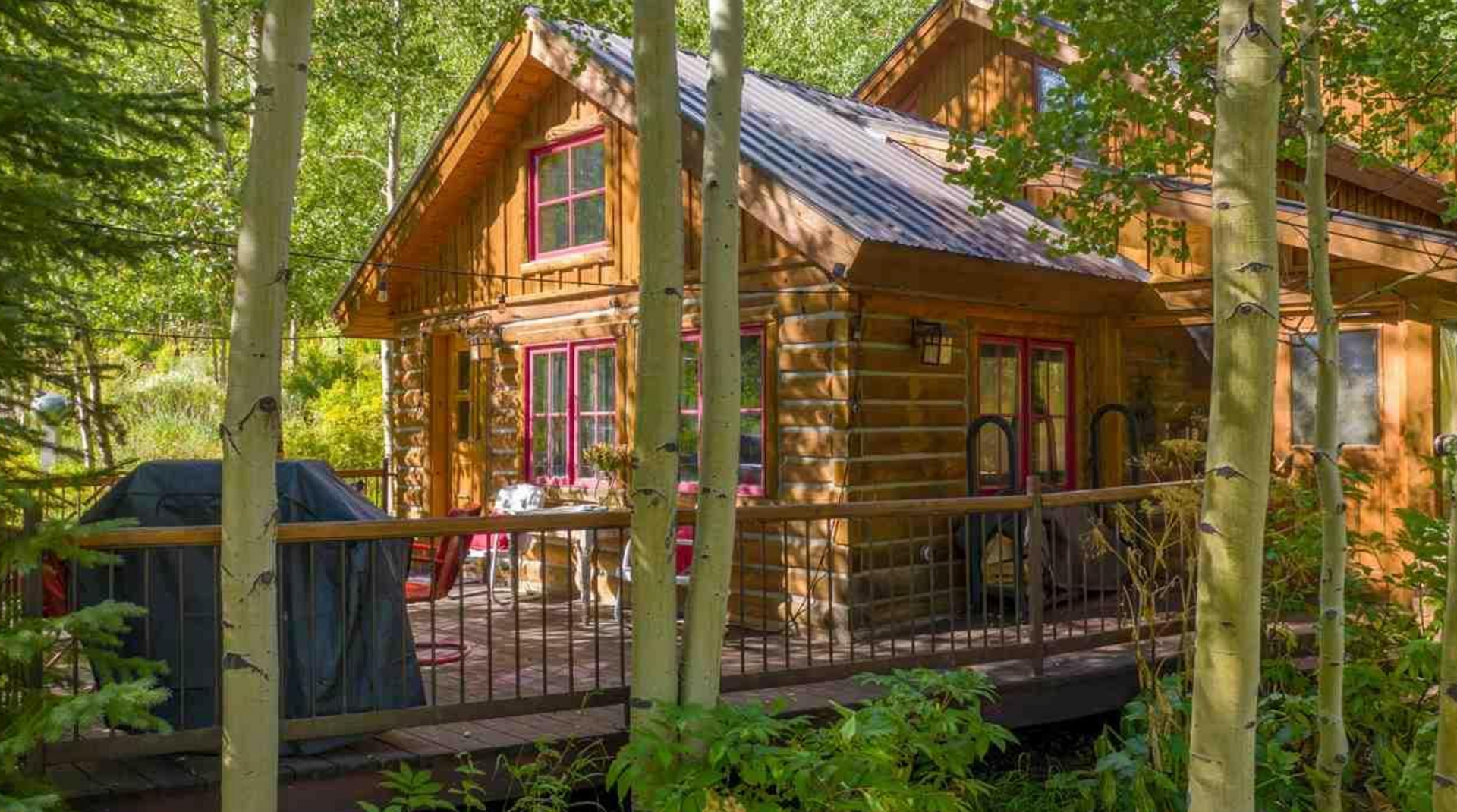 Wooden cabin with porch and metal roof in a wooded setting, surrounded by trees and greenery