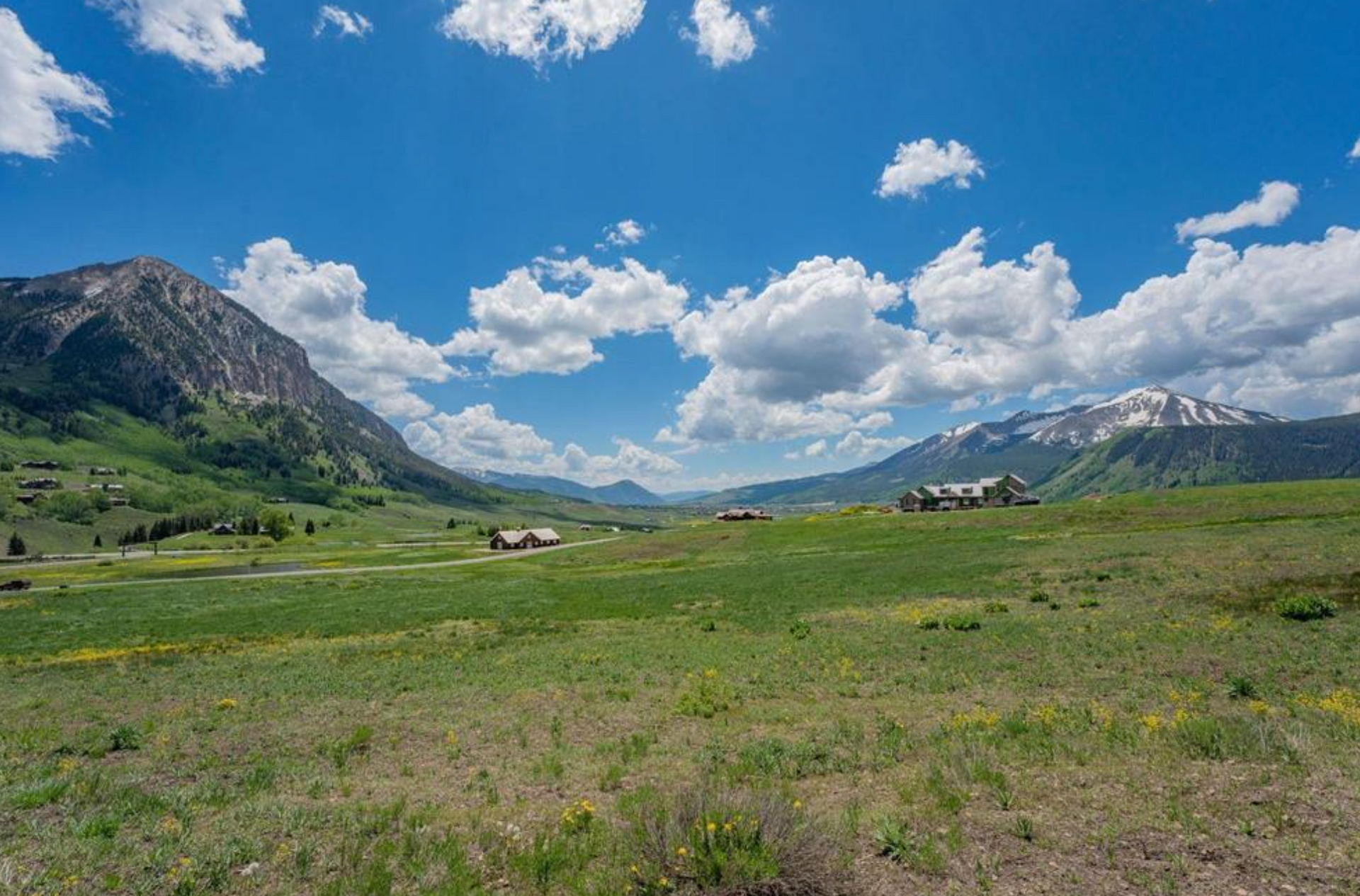 Mountain valley meadow under a bright blue sky with clouds, flanked by rocky peaks.