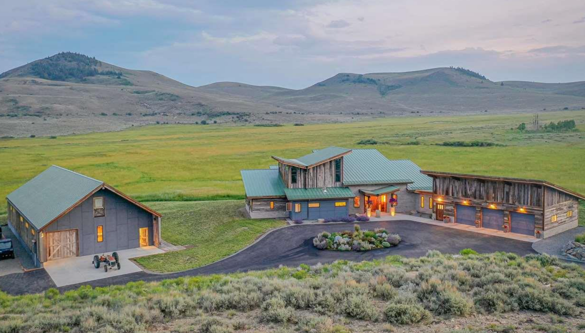 Modern ranch house and barn in a grassy valley with mountains in the background at dusk