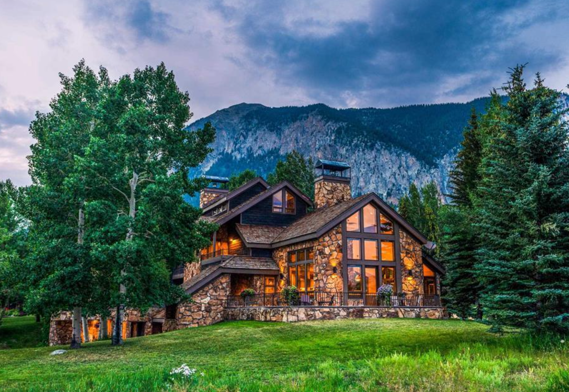 Rustic mountain cabin with warm lights, tall trees, and a dramatic sky at dusk