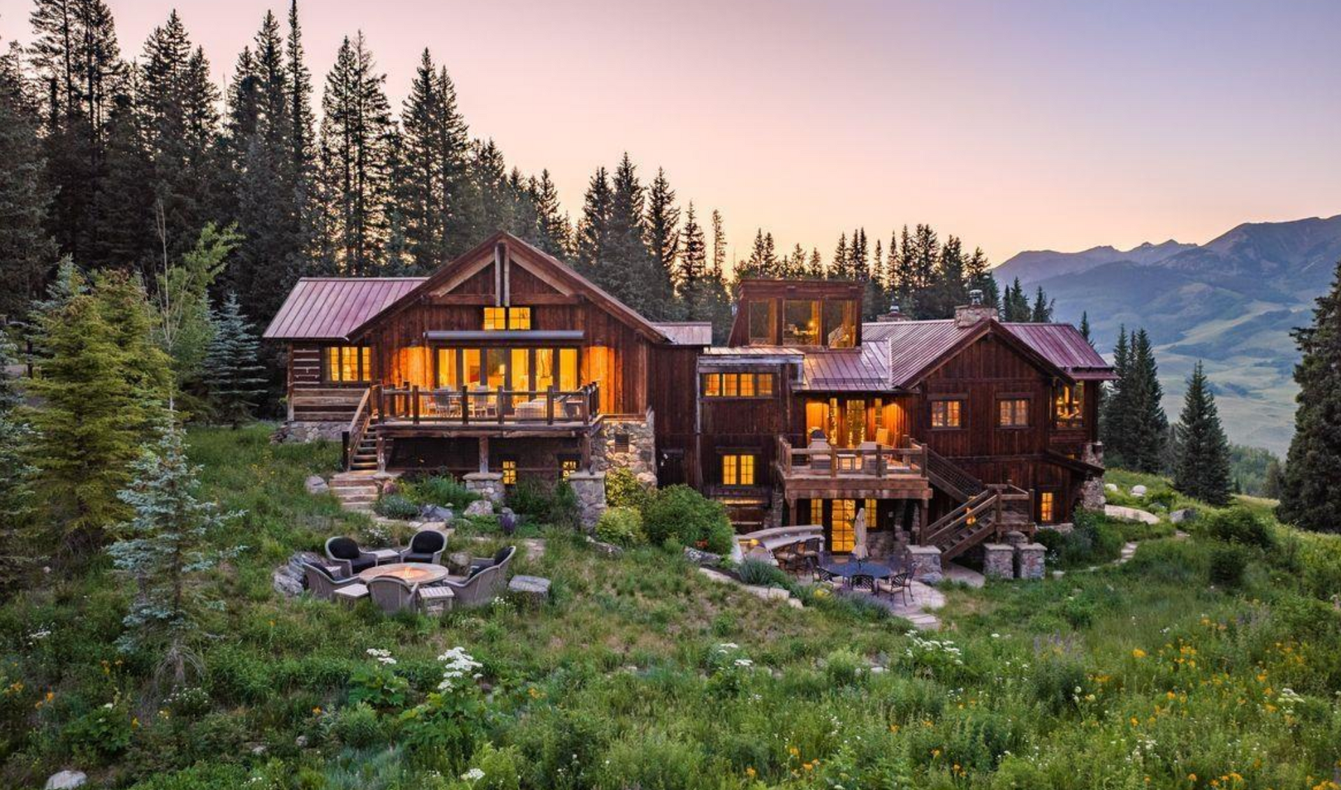 Luxury log cabins lit at dusk in a pine forest with mountains in the background