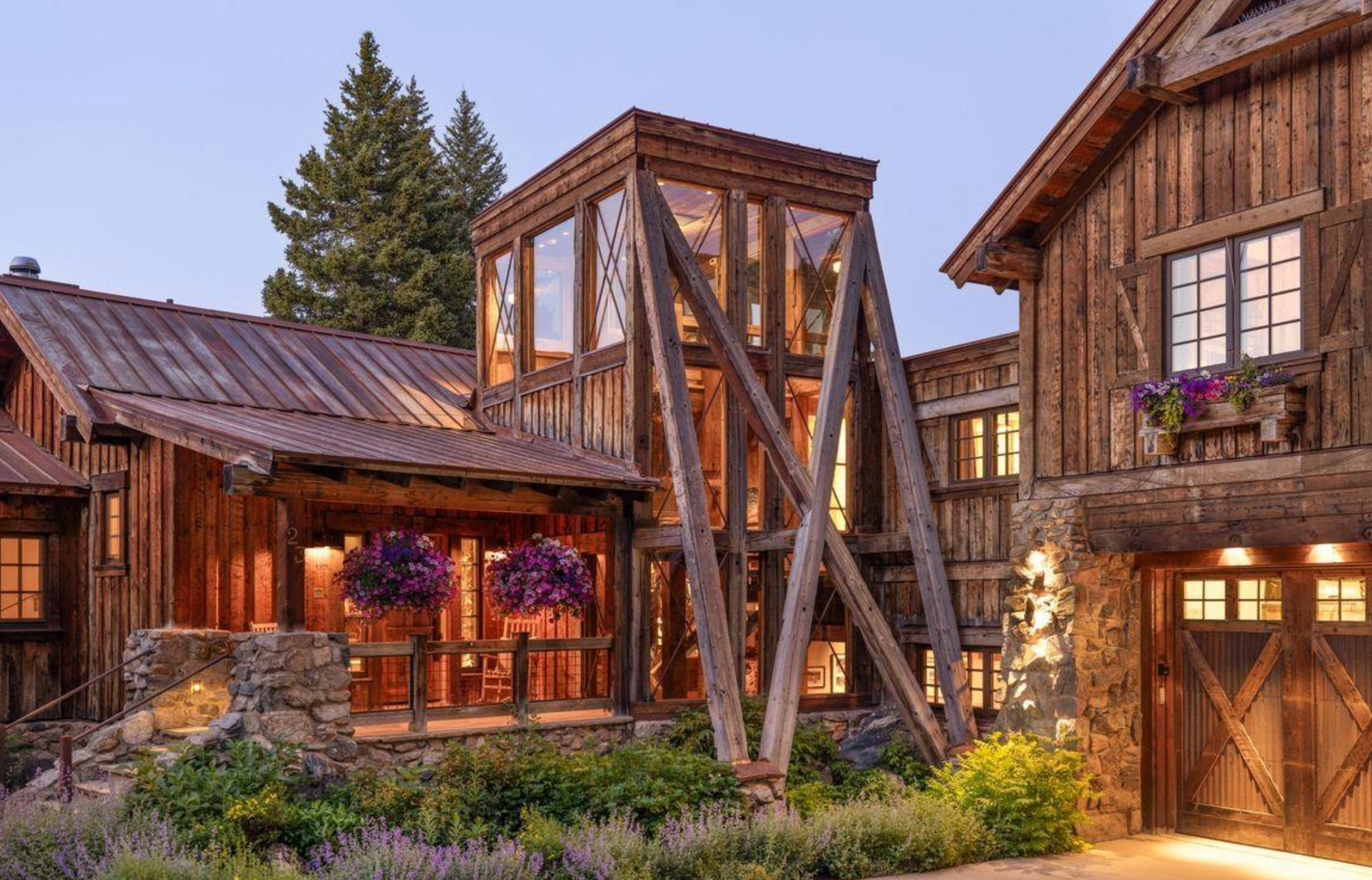 Rustic wooden lodge courtyard with a tall timber tower, stone accents, flowers, and warm evening light