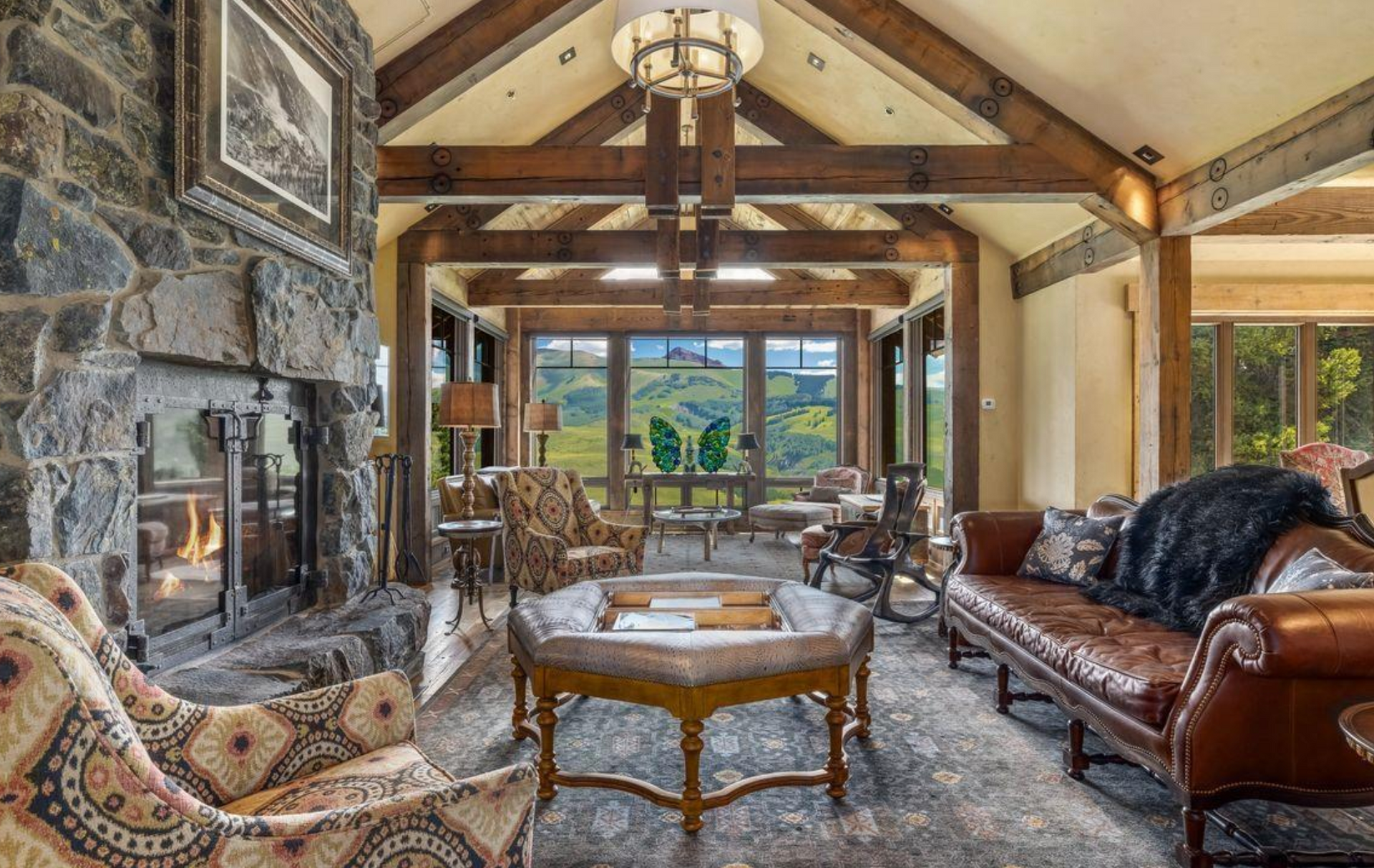 Rustic living room with stone fireplace, leather sofa, patterned chairs, and mountain view through large windows