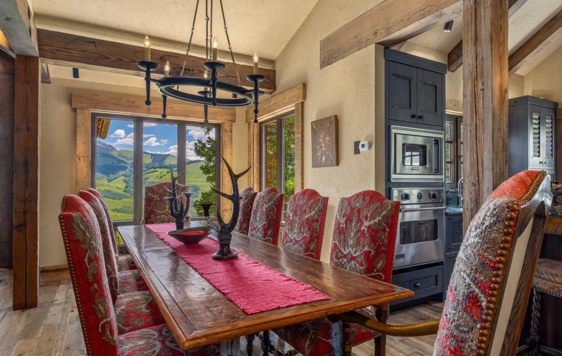 Rustic dining room with a long wooden table, red patterned chairs, and mountain-view windows