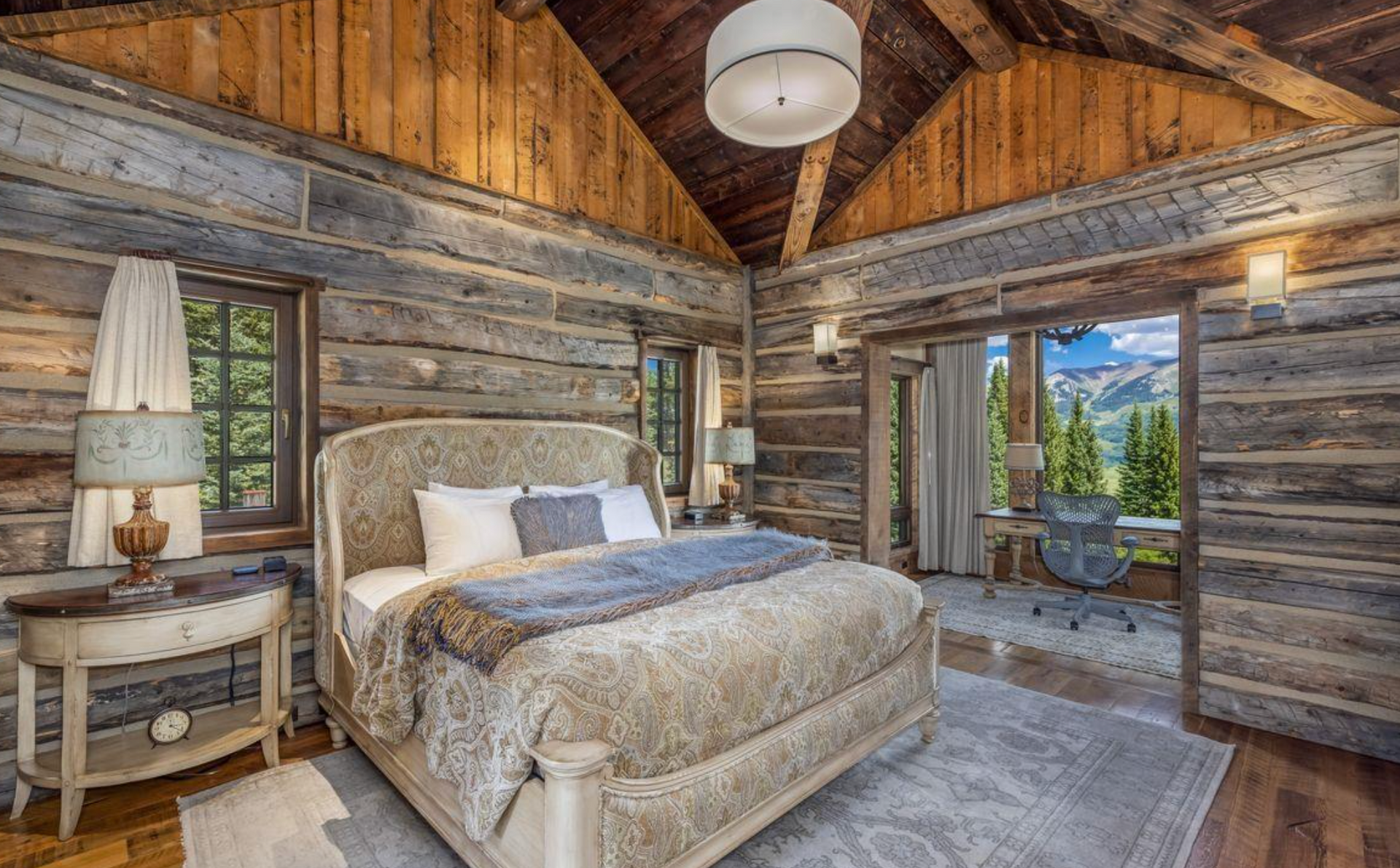 Rustic log cabin bedroom with wood ceiling, stone fireplace, bed, and open balcony view