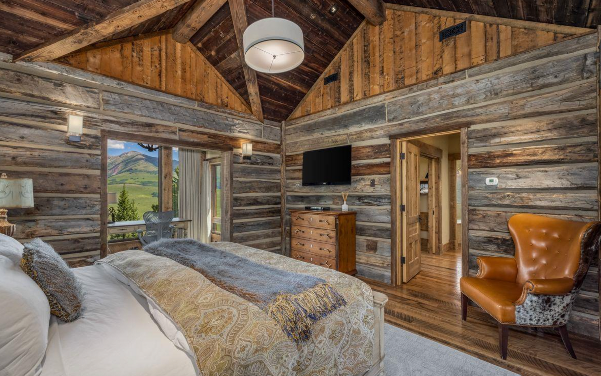 Rustic cabin bedroom with log walls, wooden ceiling, bed, TV, and mountain view through open door