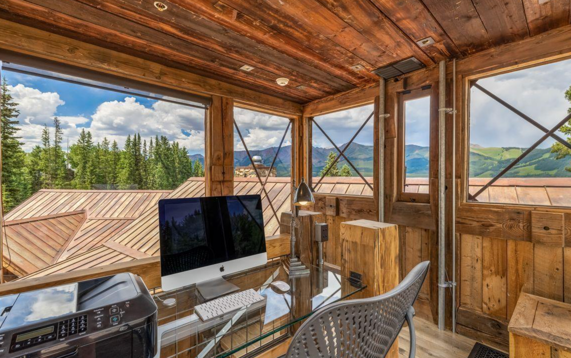 Rustic cabin workspace with a desk, computer monitor, chair, and open views of trees and mountains