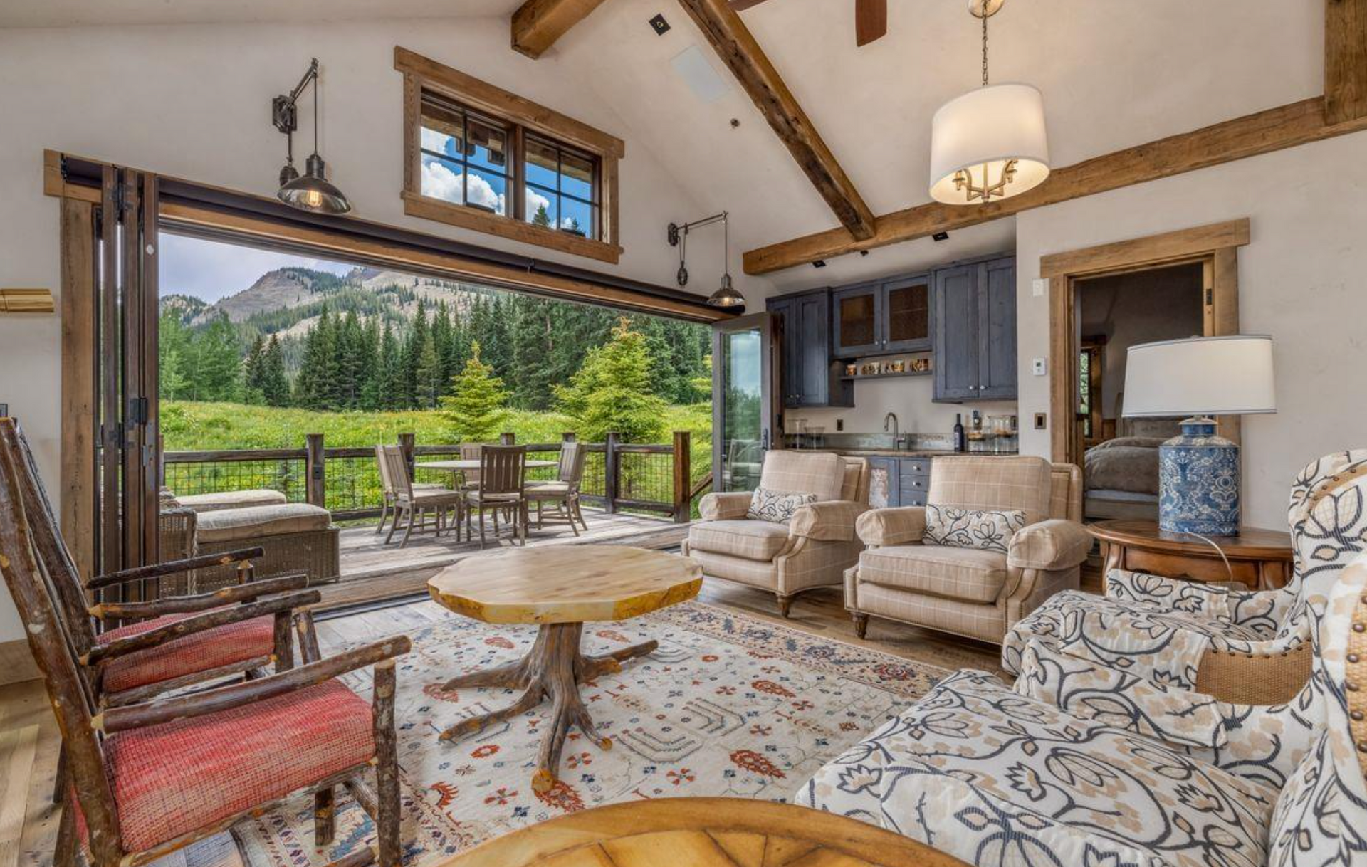 Cozy rustic living room with stone fireplace, beige sofa, patterned chairs, and large windows overlooking a mountain view