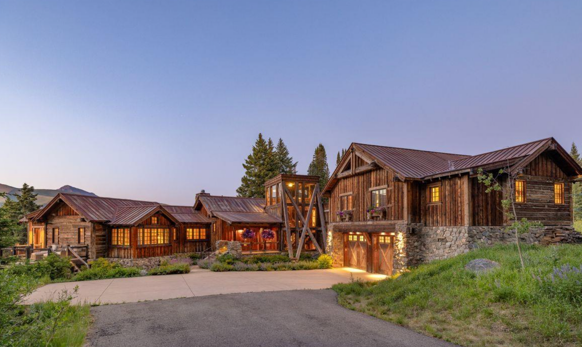 Rustic lodge-style homes at dusk with warm lit windows, stone accents, and a driveway in front