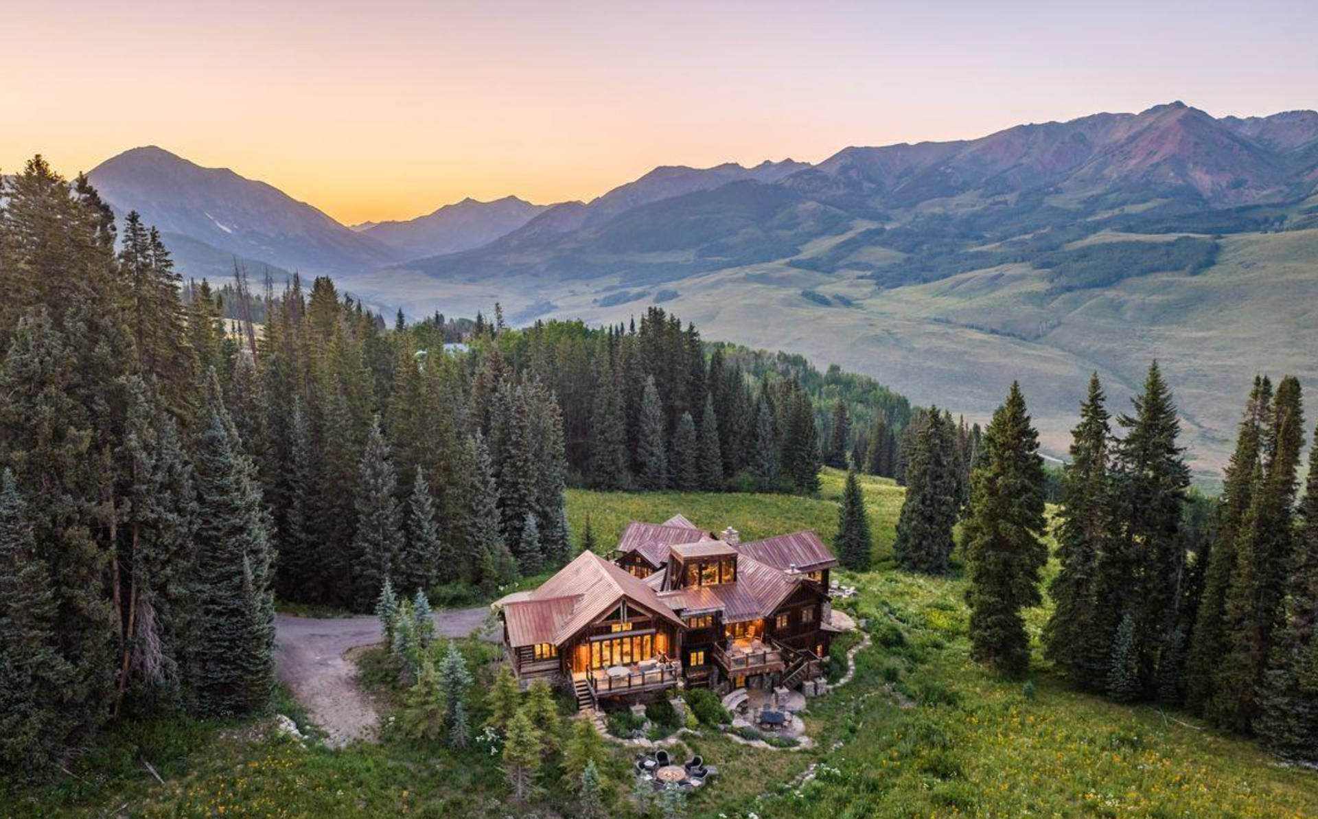 Mountain lodge among pine trees at sunset, overlooking a valley and distant mountains.