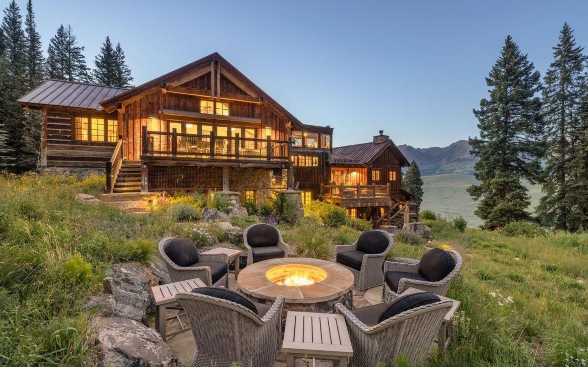 Rustic lodge at dusk with patio fire pit and Adirondack chairs in a mountain meadow