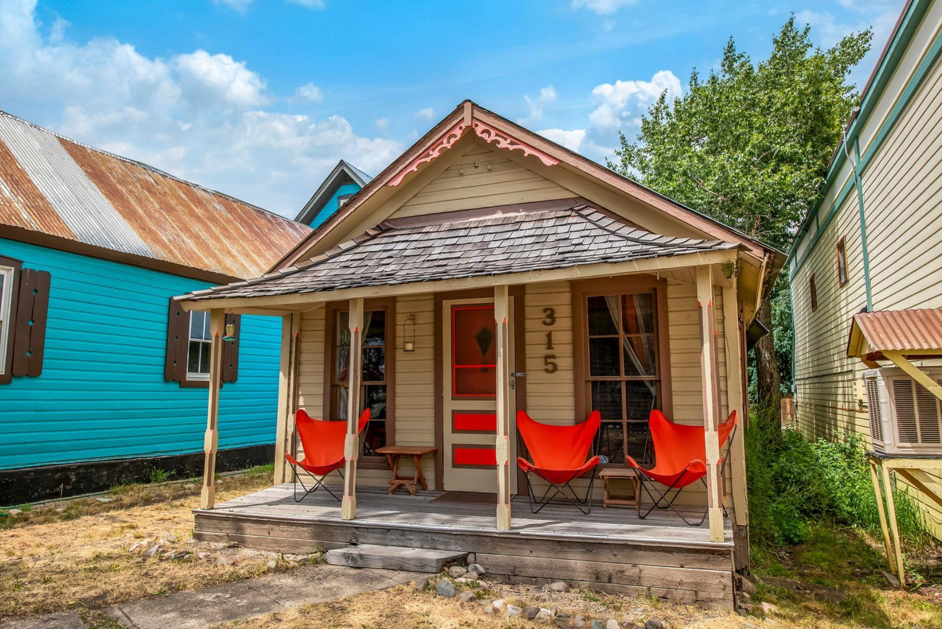 Small wooden cottage with red rocking chairs on a porch, turquoise house beside it under a blue sky