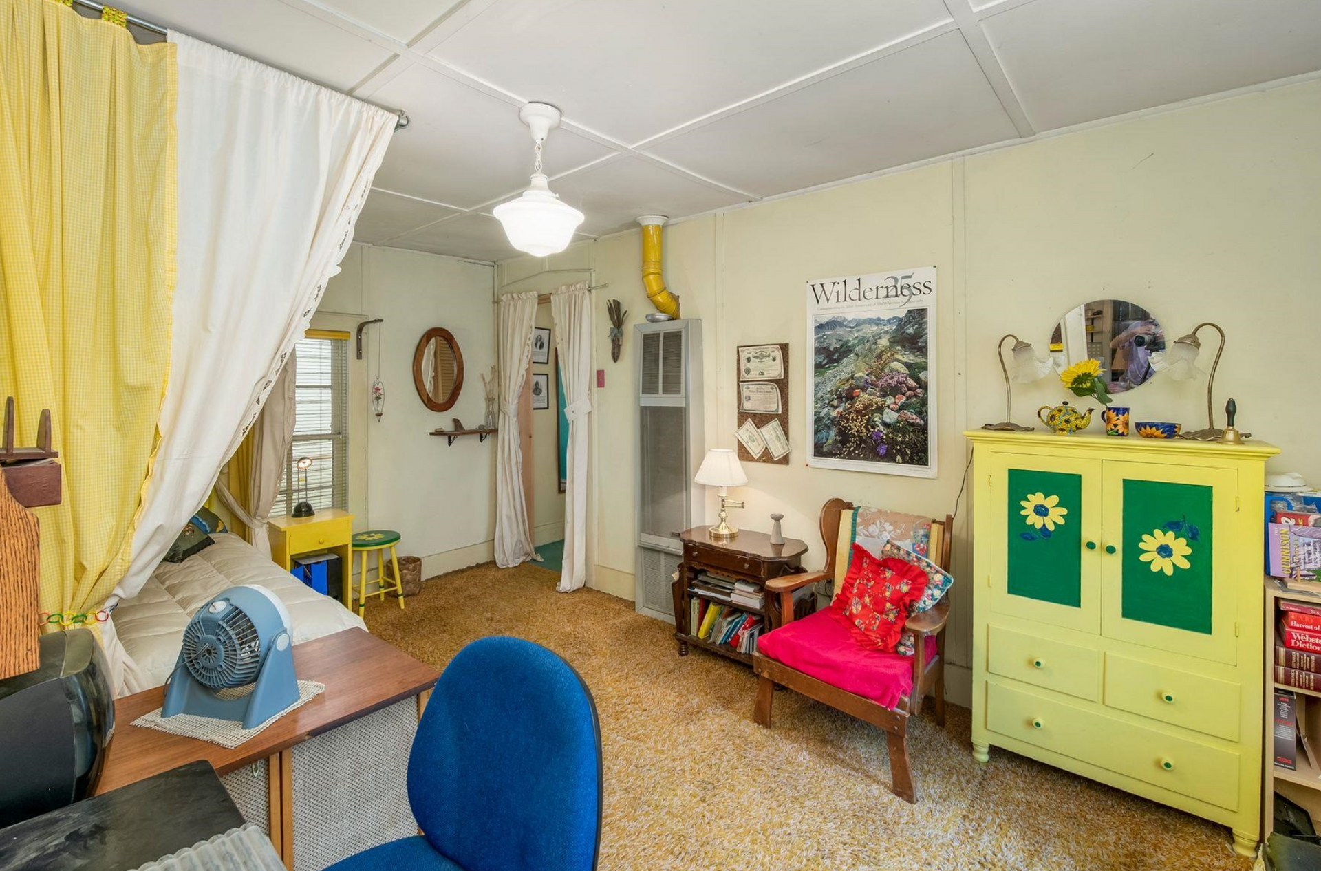 Cozy living room with yellow curtains, blue chair, red bench, and green cabinet with floral panels.