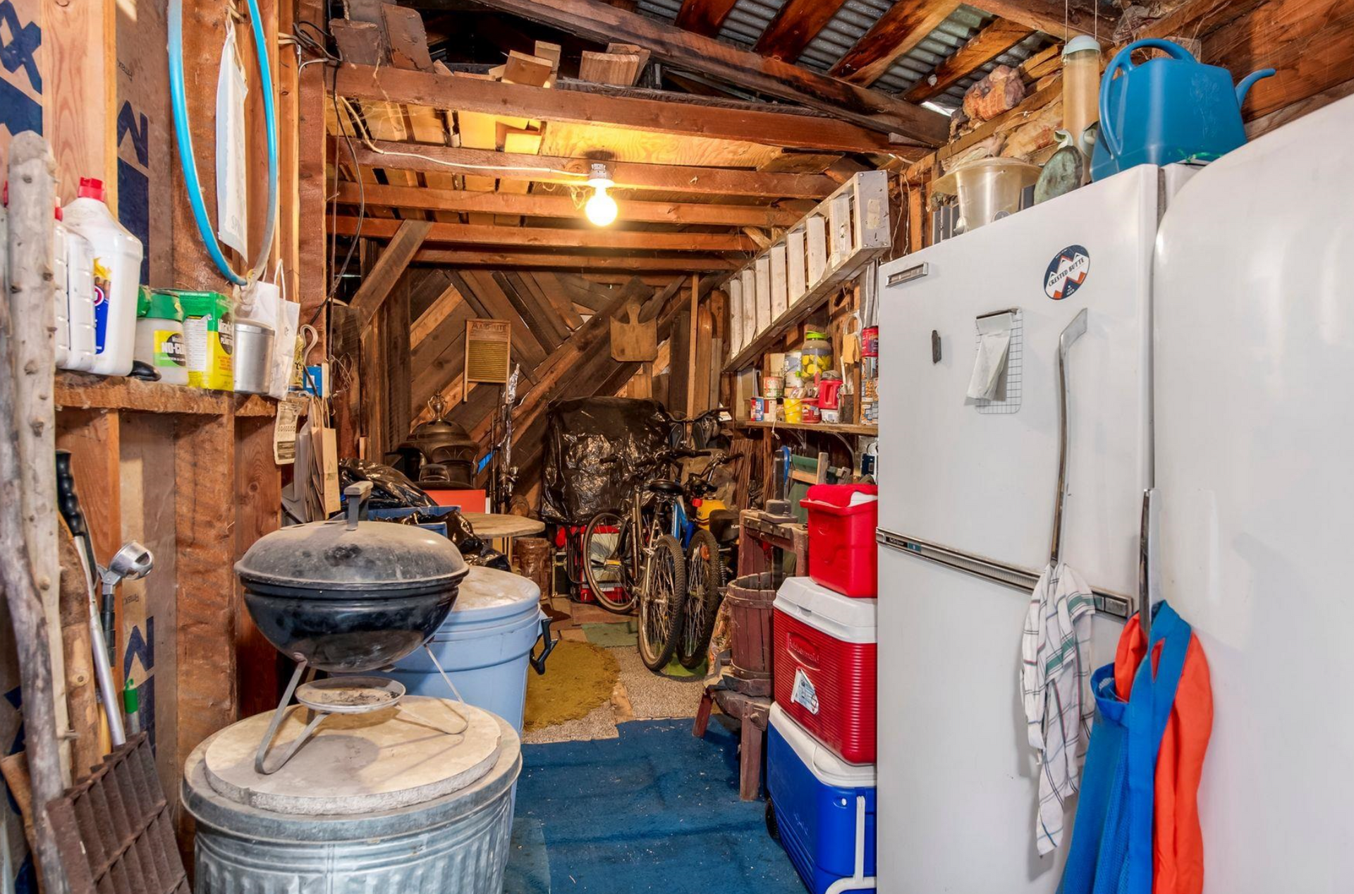 Cluttered garage workshop with a white fridge, grill, tools, and stacked supplies under exposed rafters