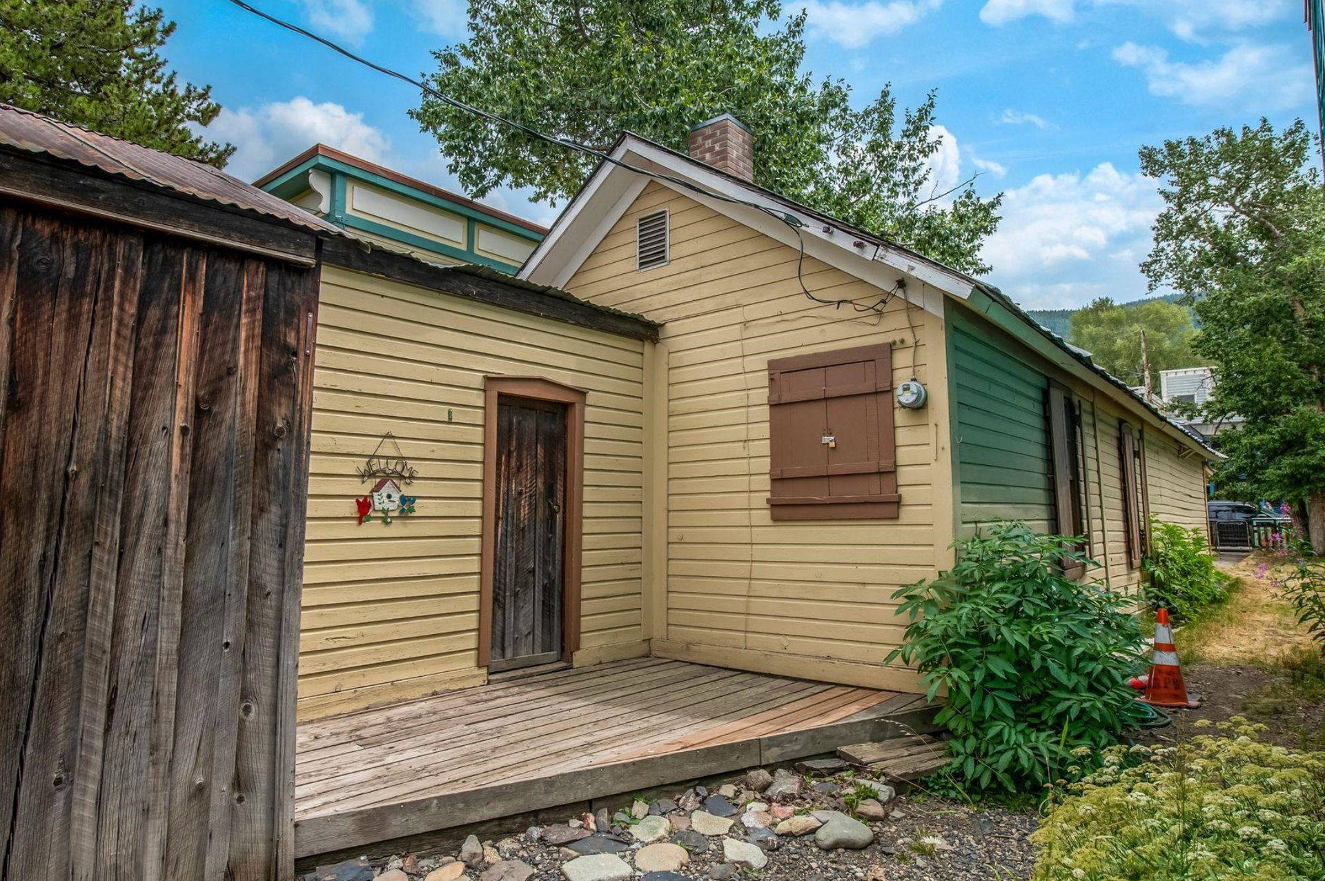 Small beige house entrance with wooden door, shuttered window, porch, and greenery beside a weathered shed