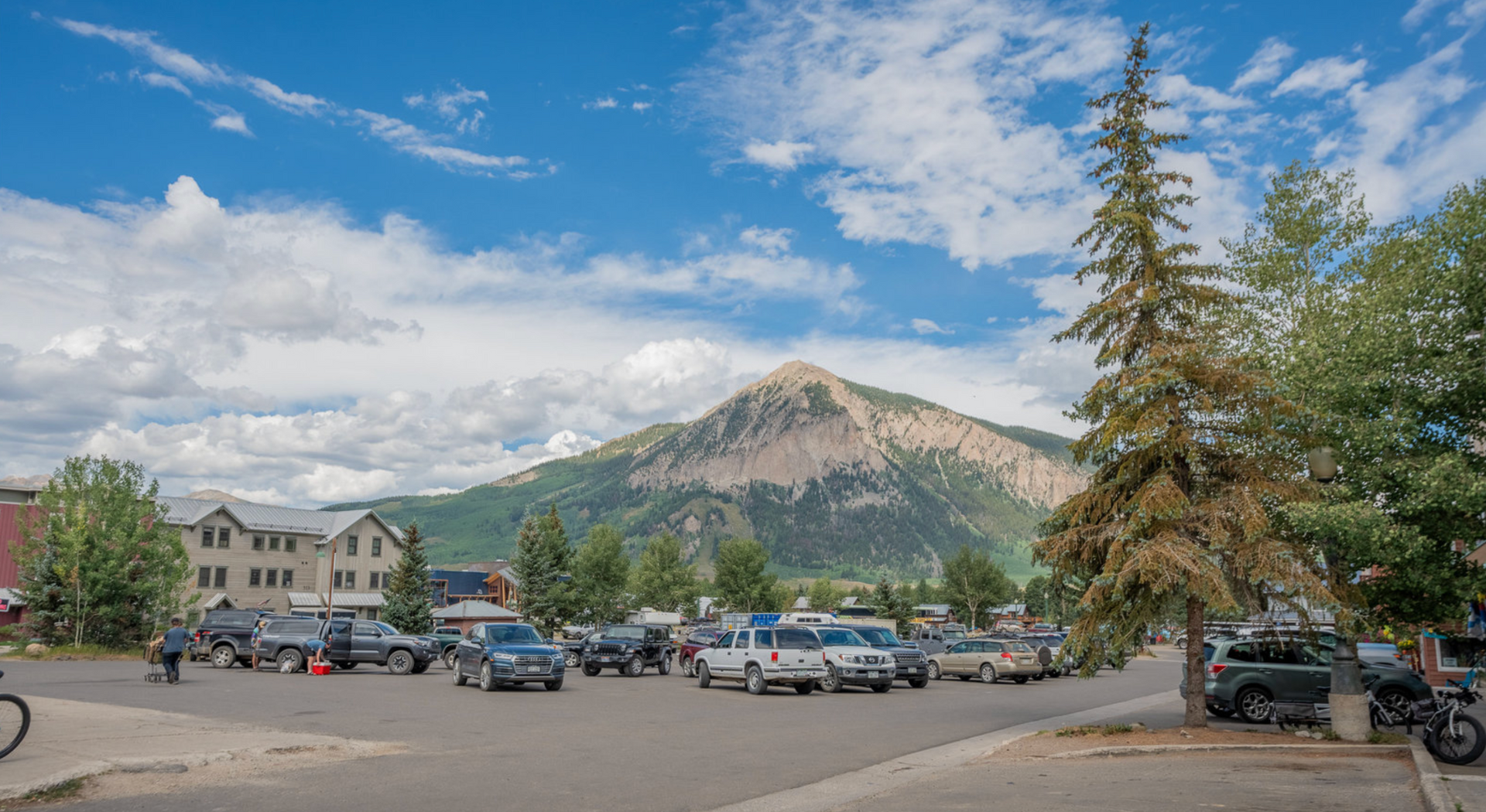 Mountain peak above a parking lot with cars, trees, and a partly cloudy blue sky