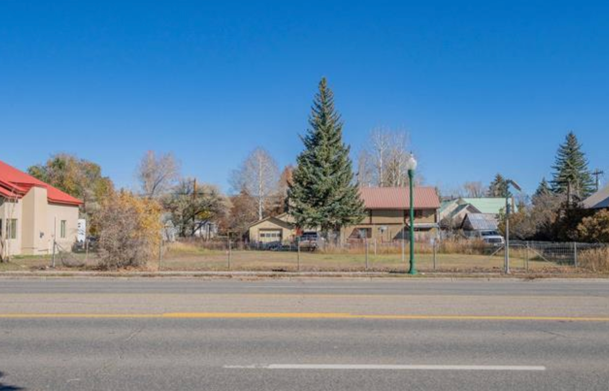 Quiet suburban street with houses, tall evergreen trees, and a clear blue sky.
